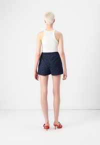 Tílko v bílé barvě, tmavě modré šortky s vysokým pasem a bílými puntíky, a červené sandály na vysokém podpatku. Modelka stojí zády proti bílému pozadí.