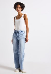 Light blue wide-leg jeans with a faded finish, paired with a white scoop-neck tank top, and white sneakers. Curly hair, hands at sides.