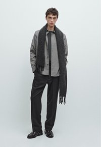 Camicia overshirt grigia, dolcevita scuro, sciarpa nera con frange, pantaloni larghi grigi e scarpe nere. Design minimalista con elementi testurizzati.