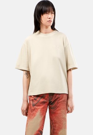 Vaalean beige oversize t-paita, lyhyet hihat, pyöreä pääntie, jossa on pieni brodeerattu kuvio rinnassa, sileä puuvillakangas.