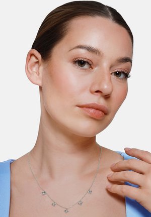 Giovane donna con pelle liscia e capelli legati indossa una collana d'argento con cinque piccoli ciondoli a forma di farfalla, vestita con un top azzurro chiaro.