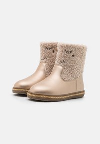 Botine metalice roz aurii cu manșete pufoase de blană bej. Prezintă detalii faciale brodate și tălpi din cauciuc brun cu cusături contrastante.