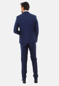 Abito blu navy con una vestibilità su misura, caratterizzato da una fessura singola sul retro. Tessuto liscio con una texture sottile, abbinato a scarpe nere lucide.