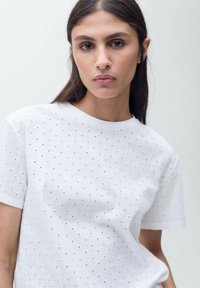 Hvid kortærmet t-shirt med et tekstureret mønster af små udskårne huller arrangeret i en diamantform. Rund halsudskæring.