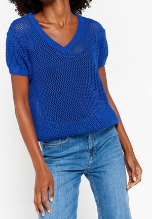 Haut en tricot bleu à manches courtes avec un décolleté en V, présentant une texture aérée et ouverte ainsi qu'un finissage côtelé, associé à un jean en denim bleu clair.
