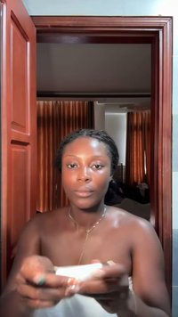 Une personne tenant un produit de soin de la peau, portant une serviette blanche. Elle a les cheveux tressés, une peau lisse et un délicat collier en or.