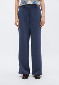 Ναυτικά παντελόνια φαρδιάς γραμμής από ομαλή ύφανση, με επίπεδη πρόσοψη, τσέπες και θηλιές για ζώνη, συνδυασμένα με σκούρα παπούτσια.