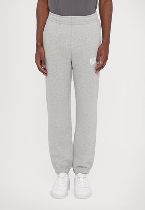 Person iført lysegrå joggingbukser med elastiske manchetter og hvide sneakers på en ensfarvet baggrund.