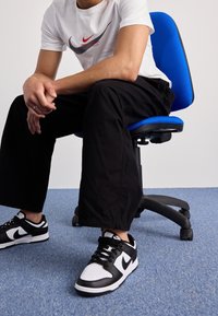 Bílý Nike tričko s červenošedým logem, černé široké kalhoty a černobílé tenisky. Sedí na modré kancelářské židli. Modrý koberec.