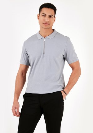 Lichtgrijze geribbelde polo shirt met een korte ritssluiting, platte kraag en korte mouwen. Draagt men met zwarte broeken, wat een getailleerd ontwerp laat zien.
