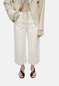 Cropped, wide-leg trousers in light cream cotton, featuring front pockets and a button closure. Paired with black flip-flops.