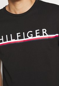 Camiseta de algodón negra con un logo blanco de "HILFIGER" y franjas horizontales rojas y azules abajo. Diseño de cuello redondo y ajuste estándar.