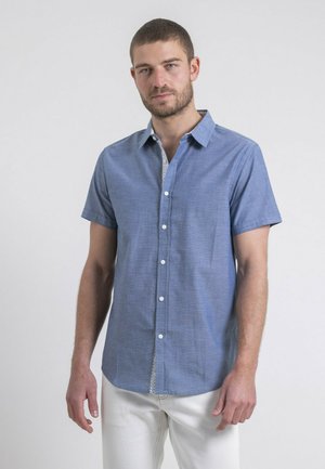 Chemise en chambray bleu à manches courtes avec un col boutonné, des boutons blancs et une patte de boutonnage à motif. Texture lisse, coupe ajustée.