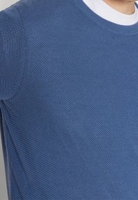 Gros plan sur une personne portant un pull en maille texturée bleu sur un t-shirt blanc à col rond, montrant les détails de l'épaule et du haut de la poitrine.