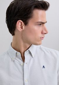 Giovane uomo rivolto a destra, indossa una camicia bianca a righe con un piccolo logo scuro di teschio e ossa incrociate sul petto.