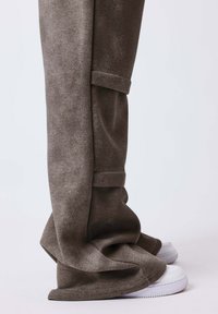 Pantalon marron à jambes larges couvrant des baskets blanches, le tissu formant des plis autour des chaussures sur un fond clair uni.