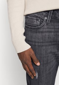 Jeans cinzentos ajustados com uma textura subtil, apresentando um detalhe de joelho desgastado, combinados com um suéter de malha canelada em creme. Mão descansando na perna da calça.