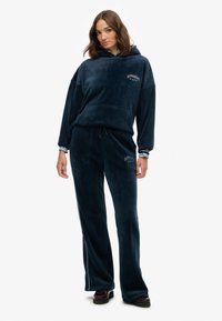 Navyblaue Velour-Jogginganzug mit Kapuzenpullover und Hose, ausgestattet mit Vordertaschen und "Superdry"-Logo in weißer Stickerei. Glatte Textur.
