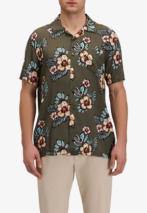 Uomo che indossa una camicia verde oliva a maniche corte con stampa floreale rossa, gialla e blu, abbinata a pantaloni beige.