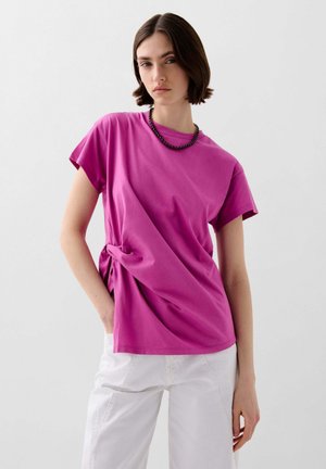 T-shirt in cotone rosa con maniche corte e dettaglio annodato sul lato, abbinato a pantaloni bianchi. Texture morbida, vestibilità rilassata, design minimal.