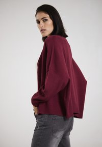 Burgunderfarbener Cardigan mit lockerer Passform, gerippten Ärmeln und Saum. Über einem hellen Top getragen, kombiniert mit grauen Jeans. Glatte Textur, entspannte Silhouette.