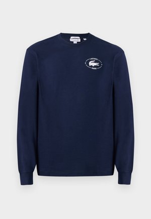 Deux personnes se tiennent côte à côte, portant des sweat-shirts bleu marine avec des logos et des jeans bleu clair, regardant dans des directions opposées sur un fond uni.