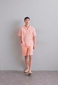 Camicetta rosa chiaro di lino a maniche corte con taschino, abbinata a pantaloni corti coordinati. L'outfit è completato da scarpe slip-on beige.