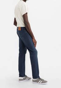 Jeans in denim blu scuro presentano un design aderente, una toppa in pelle sul retro e sono abbinati a una t-shirt chiara e a sneaker grigie.