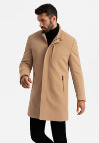 Cappotto beige, strutturato, con colletto alto, tasche laterali con zip e chiusura frontale diagonale. Indossato su un dolcevita nero e pantaloni neri.
