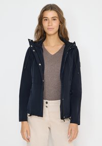 Femme portant une veste à capuche bleu marine par-dessus un pull à col en V marron et un pantalon beige, debout devant un fond blanc uni.