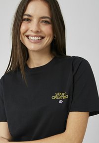 Femme souriante aux longs cheveux bruns portant un t-shirt noir avec le texte jaune "START CREATING" et une petite fleur rose en dessous.