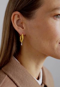 Náušnice v tvare zlata so štruktúrovaným povrchom, s visiacej priehľadnej slzičke. Nosí ich modelka so rovnými vlasmi.