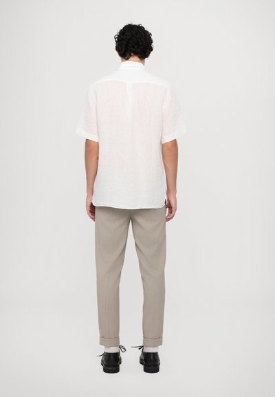 Camisa de lino blanca de manga corta con puños enrollados, combinada con pantalones gris claro y zapatos negros, vista desde la espalda contra un fondo liso.