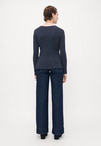 Kvinne med kort hår iført en tettsittende marineblå langermet topp og vidde mørke denimjeans, stående med ryggen til på en enkel hvit bakgrunn.