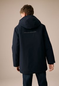 Giacca blu scuro con cappuccio e texture liscia, caratterizzata da una striscia riflettente sul retro e maniche lunghe. Indossata con jeans scuri.