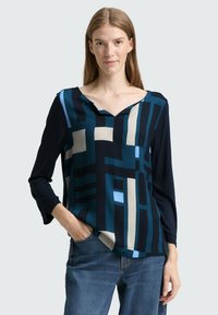 Blå och marinblå mönstrad blus med långa ärmar, V-hals och geometriska mönster i olika nyanser och texturer. Kombinerad med denimjeans.