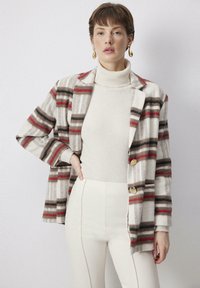 Blazer a quadri in beige, marrone e rosso, con una vestibilità rilassata, revers a dente di cane e bottoni dorati. Indossato sopra un dolcevita crema.