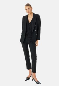 Completo nero su misura con giacca doppiopetto, gilet aderente e pantaloni slim, abbinato a décolleté nere a punta.