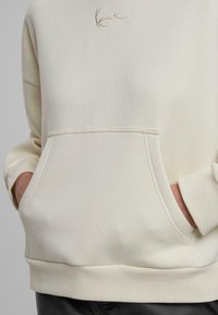 Helle Hoodie in Cremefarbe mit einer vorderen Tasche, gerippten Bündchen und Saum. Mit dezentem, kursiv gesticktem Schriftzug über der Tasche. Glatte Textur.
