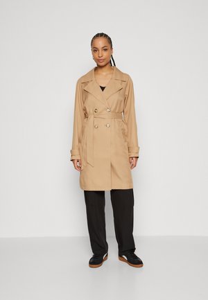 Trench coat beje în design dublu în culoare cu un cordon, având nasturi mari și o croială relaxată. Purtați peste pantaloni și pantofi negri.