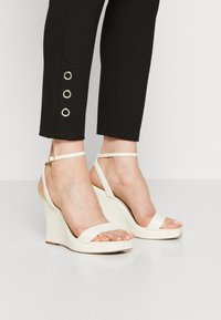 Weiße Keilsandalen mit glatter Oberfläche, Design mit zwei Riemen, verstellbarem Knöchelriemen und hohem, geschwungenem Absatz. Schwarze Hose mit silbernen Ösen.