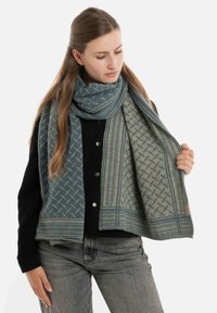 Geweven sjaal in blauw en beige met geometrische patronen, franjes, en een zachte textuur, gedragen over een zwart sweater.