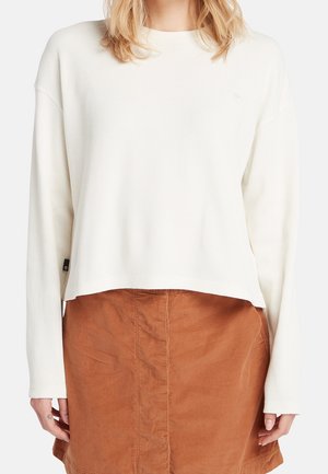 Žena vynášející bílý žebrovaný svetr s dlouhým rukávem v kombinaci s hnědou manšestrovou sukní s vysokým pasem na neutrálním pozadí.