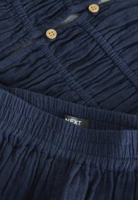 Tissu texturé bleu marine avec des détails froncés et deux boutons en bois ; comprend une étiquette visible portant la mention "NEXT." Convient pour une tenue décontractée.