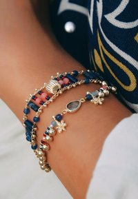 Bracelet multi-brins avec des perles bleu foncé, des accents roses et rouges, des éléments en métal doré, un charm en forme de coquillage et des détails en forme de fleur.