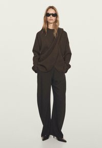 Frau trägt einen übergroßen, dunkelbraunen, drapierten Pullover, weite schwarze Hosen, schwarze spitze High Heels und schwarze Sonnenbrille, steht vor einem schlichten Hintergrund.