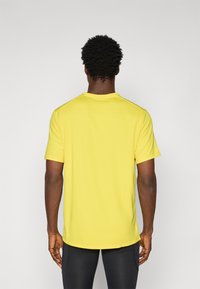 Chemise jaune à manches courtes avec un col rond et une coupe décontractée. Le tissu semble lisse sans motifs ni accents visibles. L'arrière-plan est uni.