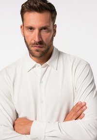 Witte poloshirt met lange mouwen, van zachte stof, met een geknoopte kraag en een subtiel logo op de borst. Het model poseert met gekruiste armen.