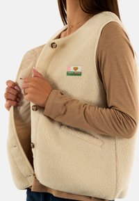 Gilet in pile color crema con una superficie texturizzata, dotato di bottoni e una toppa con la scritta "ONLY VINTAGE", indossato sopra una maglietta beige a maniche lunghe.