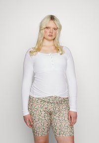 Blusa branca de mangas compridas em ribana com um decote abotoado, combinada com calções verdes estampados com flores em tons de rosa e branco.